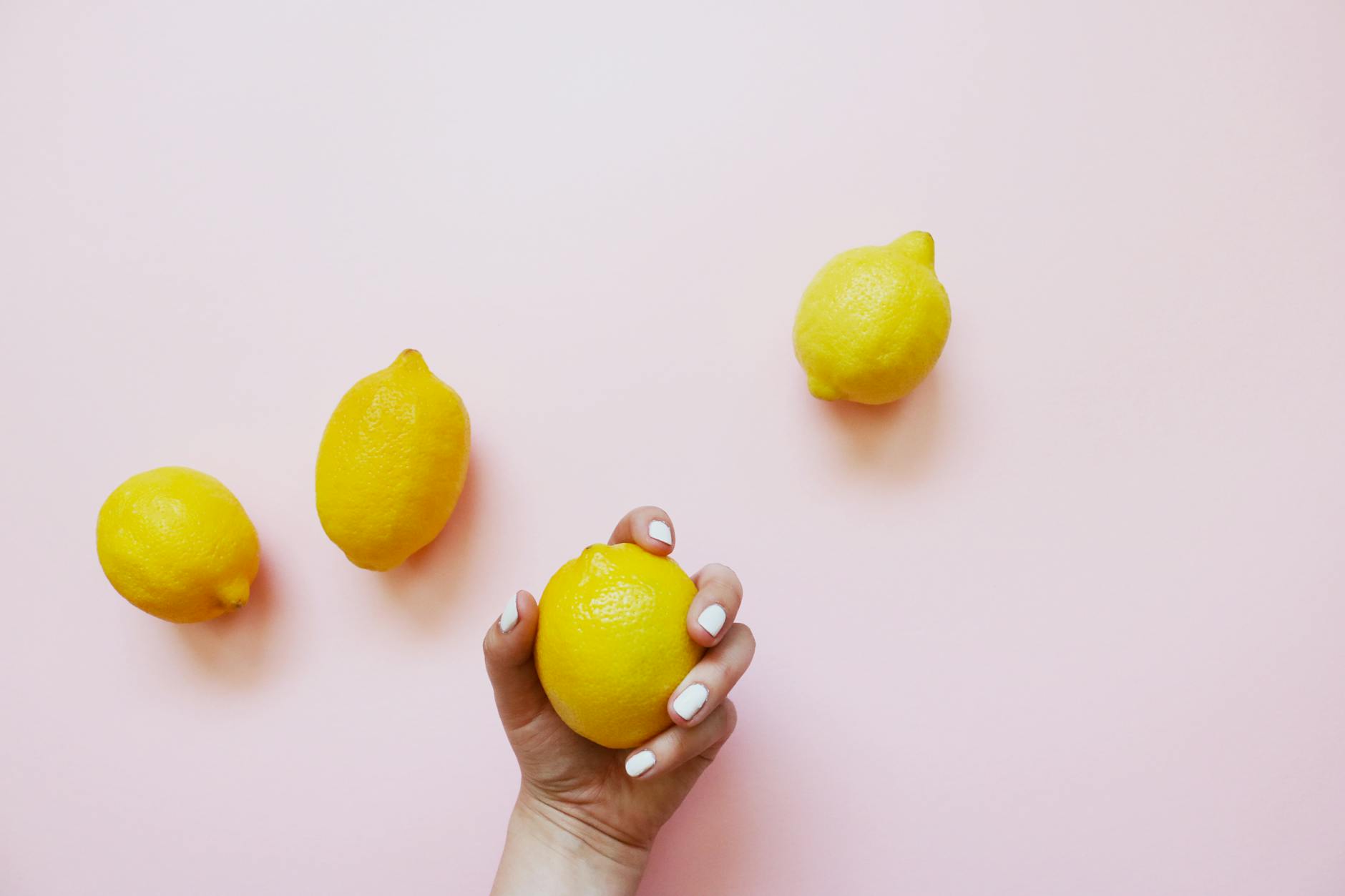 A hand with white nails holding a lemon on a soft pink background, surrounded by lemons