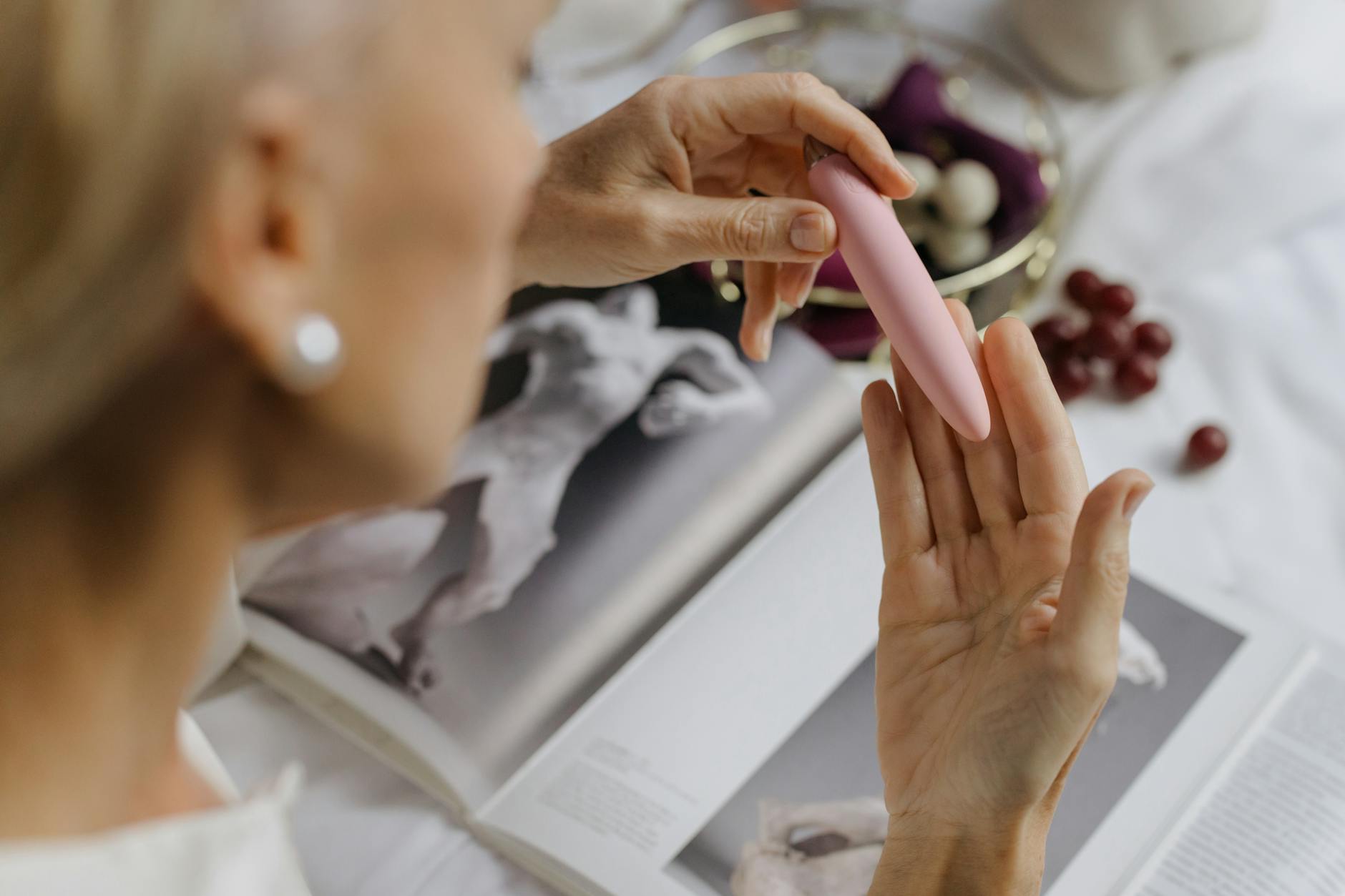 A close-up of a woman holding a pink vibrator, with art books in the background, representing intimate self-exploration.